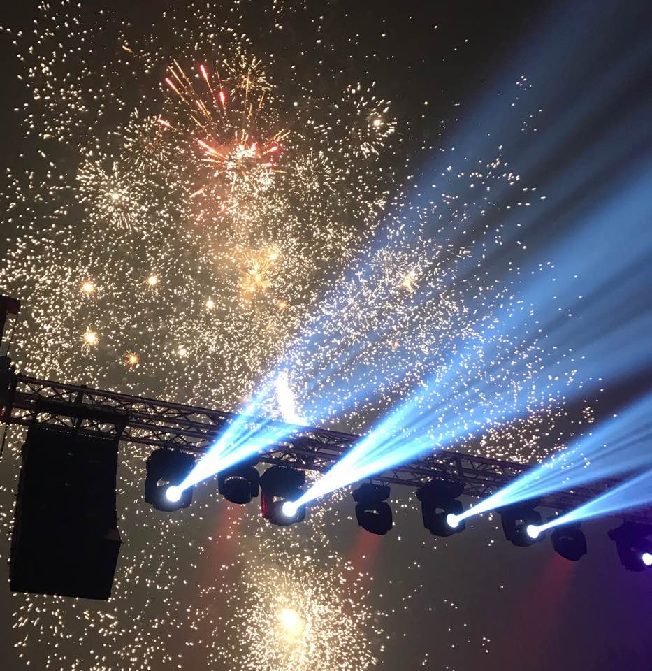 Fuochi d'artificio scintillanti sopra un palco illuminato da riflettori blu durante un festival