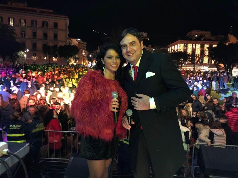 Christian Apadula e un'artista di palco, Shasa, sorridono davanti a una folla durante un evento serale in piazza.